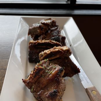 Woori - Kalbi - A rectangular white plate contains multiple pieces of grilled beef short ribs (kalbi) with a caramelized glaze, garnished with chopped herbs. The plate is positioned on a wooden surface near a window, with an outdoor scene visible through the glass, including a person and a bicycle. The image emphasizes the presentation of the Korean dish in a casual dining setting. - Korean Near Me - Arlington, Massachusetts