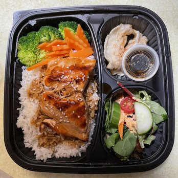 Eggs & Thai Café - Salmon lunch. - A black plastic bento box containing rice topped with grilled chicken, steamed broccoli, carrots, a side of dumplings, a small salad, and a dipping sauce. - Thai Near Me - Dedham, Massachusetts