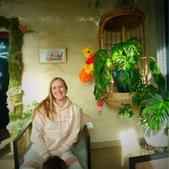 Florfe Cafe - A woman with long blonde hair, wearing a light-colored hoodie, sits smiling in a cafe decorated with plants and Winnie the Pooh plush toys. - Cafes Near Me - Salem, Oregon