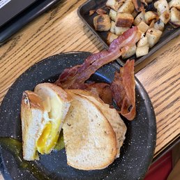 Better with Cheddar - The image shows a breakfast plate with a fried egg sandwich on toasted bread, with the yolk slightly runny. Accompanying the sandwich are two strips of crispy bacon and a side of roasted potatoes, which are in a small metal tray. The meal is placed on a wooden table, with part of a laptop visible in the background.