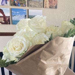 A bouquet of white roses with lush petals and green leaves is wrapped in brown paper. The bouquet is placed indoors, with a wall in the background decorated with a collage of travel photographs featuring natural landscapes and landmarks.