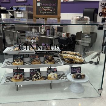 Cinnaholic - The dessert choices. - A glass display case contains an assortment of cinnamon rolls, cookies, and baked desserts, each labeled with small signs indicating flavors like Oreo, strawberry, and baby buns. The case is situated on a white counter, with additional baked goods and promotional signs nearby. The background includes a purple wall, a chalkboard, and bakery equipment, indicating an indoor bakery setting. - Bakeries Near Me - Tucson, Arizona