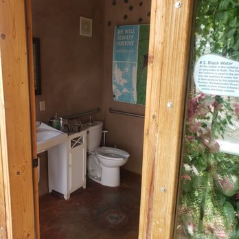 Earthship Biotecture - You can use this with your admission price - A small bathroom features a white toilet, a white sink with a small cabinet, and a window with greenery outside. The walls are plain, and there is a poster on the wall with survival information. The floor appears to be concrete, and there are metal grab bars near the toilet. The bathroom is part of an eco-friendly or sustainable building, as indicated by the wooden framing and the poster. - General Contractors Near Me - Tres Piedras, New Mexico