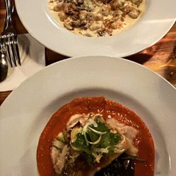 Gnocchi - The image shows two plates of pasta dishes on a wooden table in a restaurant setting. The top plate contains pasta with a creamy sauce, mushrooms, and possibly walnuts and crispy onions. The bottom plate features pasta with a tomato-based sauce, garnished with greens and herbs. Silverware and a napkin are visible nearby, indicating a dining environment.
