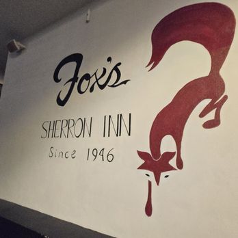 Fox’s Lounge - The image shows a wall with a painted logo and text for Jox's Sherron Inn, established in 1946. The logo includes the name 'Jox's' in a stylized script, with 'SHERRON INN' and 'Since 1946' written below. To the right, there is a large, stylized illustration of a fox with a flowing tail, depicted in red, with a drip of red liquid from its mouth, possibly symbolizing a signature or branding motif. The setting appears to be indoors, with part of a striped awning visible in the background. - Dive Bars Near Me - Miami, Florida
