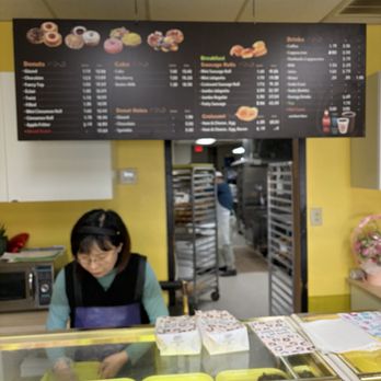 Young Donut - A bakery counter shows a woman working behind the glass display case filled with baked goods. Above her, a large menu board lists items such as donuts, cakes, and beverages. The background includes a kitchen area with a person working and bakery racks. The setting appears to be a bakery or donut shop. - Donuts Near Me - Irving, Texas