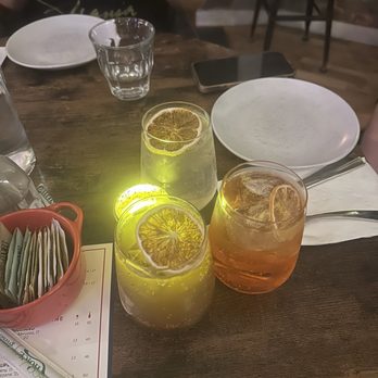Vineapple - Three beverages with lemon slices are on a wooden table, accompanied by empty plates, a glass of water, and a smartphone, in a restaurant setting. - Italian Near Me - Brooklyn, New York