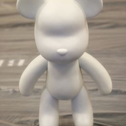 A smooth, white bear-shaped figurine with rounded ears, limbs, and a body, but lacking facial features or other details. It stands upright on a wooden surface with a blurred background, and appears to be made of a matte material, possibly ceramic or plastic.