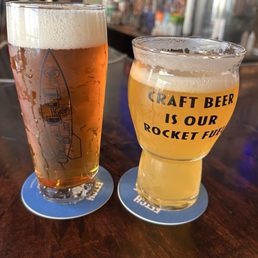 Two glasses of craft beer are positioned on a dark wooden bar counter. The taller glass contains a golden amber beer with a foamy head, while the shorter glass holds a pale yellow beer with a slight haze. The shorter glass features the phrase "Craft Beer is Our Rocket Fuel" printed in black. The background shows a blurred bar setting with various items and decorations, indicating a lively environment.