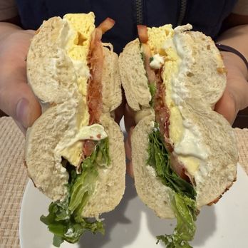 Big Apple Bagels - BLT with egg on an onion bagel - A person holds a halved BLT sandwich with egg, bacon, lettuce, and possibly mayonnaise on an onion bagel. The sandwich is cut in half, showing the layers of ingredients inside. The person is wearing a dark jacket, and the background includes a woven placemat and a white plate. The image emphasizes the sandwich's ingredients and presentation. - Bagels Near Me - Aurora, Illinois