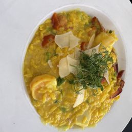 Seafood Risotto - A seafood risotto is presented on a white, round plate, featuring yellow rice with visible pieces of seafood, garnished with fresh herbs, cheese shavings, and green vegetables. The dish appears creamy and well-prepared, suitable for a Mediterranean restaurant setting.