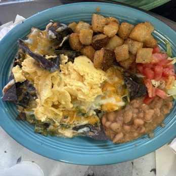The Chancla ABQ - A breakfast plate served on a blue oval dish includes scrambled eggs with melted cheese, crispy roasted potatoes, baked beans, and a side of chopped fresh tomatoes and lettuce. The meal is arranged with the eggs and potatoes occupying most of the plate, and the tomato salad on the side. The plate is placed on a table with napkins and utensils nearby. - Breakfast & Brunch Near Me - Albuquerque, New Mexico