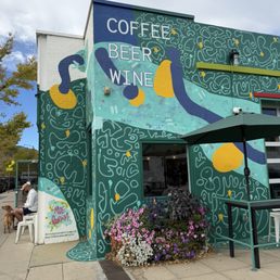 The image shows the exterior wall of a building decorated with a vibrant mural that includes abstract shapes and patterns. The mural prominently displays the words "COFFEE," "BEER," and "WINE" in large letters. There is a small outdoor seating area with a green umbrella and a flower bed with pink, purple, and white flowers. A person is sitting on a chair with a dog nearby, and the sky is partly cloudy.