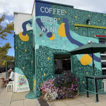 Creature Comforts Cafe - The image shows the exterior wall of a building decorated with a vibrant mural that includes abstract shapes and patterns. The mural prominently displays the words "COFFEE," "BEER," and "WINE" in large letters. There is a small outdoor seating area with a green umbrella and a flower bed with pink, purple, and white flowers. A person is sitting on a chair with a dog nearby, and the sky is partly cloudy. - Coffee & Tea Near Me - Boulder, Colorado