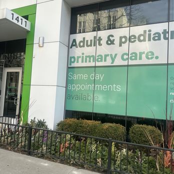 Rush South Loop - Exterior. - The image shows the exterior of a medical clinic with a large glass window displaying text about adult and pediatric primary care services, including same-day appointments. The building has a white facade with green accents, and the address number 1411 is visible near the entrance. There is a small garden with bushes and flowers in front of the window, bordered by a black metal fence. The sidewalk runs along the front of the building. - Doctors Near Me - Chicago, Illinois