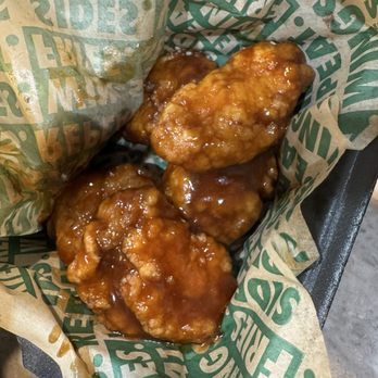 Wingstop - Hawaiann boneless wings - Four glazed chicken wings are placed in a paper-lined basket, with a sticky sauce coating them. The basket is on a dark surface. - Chicken Wings Near Me - Fairfax, Virginia