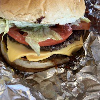 Five Guys - Bacon Cheeseburger (all the way) - A bacon cheeseburger features a sesame seed bun, a beef patty topped with melted cheese, crispy bacon, fresh lettuce, and a slice of tomato. The burger is partially wrapped in crinkled aluminum foil, indicating it is ready for serving or takeout. - Burgers Near Me - Santa Fe, New Mexico