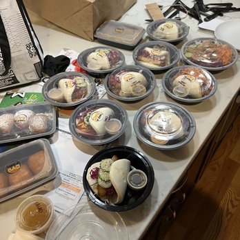 Take a Bao Eats - Ridiculous amount of baos! Crispy Spicy Aioli Chicken Bao, pork belly bao, Teriyaki Chicken Bao. - Several takeout bao meals in plastic containers with clear lids are arranged on a kitchen countertop. The bao include different fillings, and some are accompanied by small containers of sauces. Additional side dishes and salads are also present, with some containers open or partially covered. The background shows a paper bag, utensils, and miscellaneous kitchen items, indicating a recent meal pickup or delivery. - Asian Fusion Near Me - Chicago, Illinois
