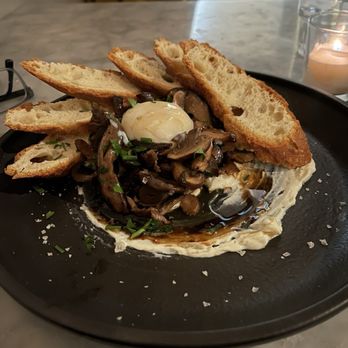 Root & Flower - Braised mushrooms with egg - A black plate holds a serving of sautéed mushrooms garnished with chopped chives, topped with a poached egg. The dish is accompanied by several slices of toasted bread arranged around the mushrooms. The plate is placed on a marble table, with a candle and glasses visible in the background, indicating a dining setting. - Wine Bars Near Me - Vail, Colorado