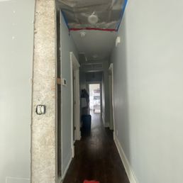 Before repair work - A hallway with light gray walls and dark wooden flooring, featuring exposed ductwork and insulation on the ceiling. Construction materials and tools are visible, indicating ongoing renovation work. The hallway leads to a room with natural light and furniture visible in the background.