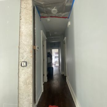 Skinner and Son - Before repair work - A hallway with light gray walls and dark wooden flooring, featuring exposed ductwork and insulation on the ceiling. Construction materials and tools are visible, indicating ongoing renovation work. The hallway leads to a room with natural light and furniture visible in the background. - Painters Near Me