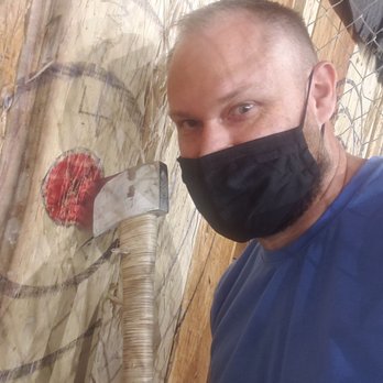 Craft Axe Throwing- Springfield - A man with short hair and a beard, wearing a black face mask and a blue shirt, is taking a selfie at a wood chopping station. The background includes a wooden wall and a chain-link fence. An axe is embedded in a log on the chopping block, which is positioned vertically. The log appears to be part of a wood chopping activity or station. - Axe Throwing Near Me - Springfield, Missouri