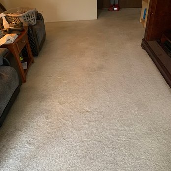 A-Plus Cleaning Service - Blood stain gone! - A living room with a beige carpet, a dark sofa, a small wooden side table, and household items including a laundry basket and a vacuum cleaner. - Carpet Cleaning Near Me - Chicago, Illinois