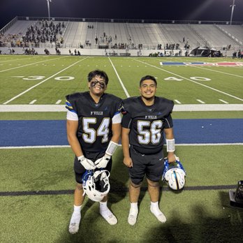 The Pfield - These guys rock and worked hard! - Two football players in black jerseys with the numbers 54 and 56 stand on a football field at night, holding their helmets and smiling. The field has white yard lines and a blue end zone. In the background, there are bleachers with some spectators and bright stadium lights illuminating the scene. - Stadiums & Arenas Near Me - Pflugerville, Texas