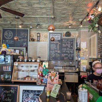 OK Cafe - The image shows the interior of a coffee shop with a decorative ceiling, ceiling fans, and warm lighting. A barista wearing glasses and a pink mask is working behind the counter, which has coffee cups, a green espresso machine, and various supplies. The background includes a chalkboard menu listing coffee options, a display case with baked goods, and shelves with decorations and supplies. The decor includes patterned wallpaper, hanging lights, and plush toys. - Coffee & Tea Near Me - Astoria, New York