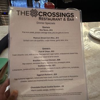 The Crossings - Menu - The menu from The Crossings Restaurant & Bar features dinner specials such as starters, entrees, and desserts. Items include Dos Tacos, Mexican Street Corn Ribs, Fish N' Chips, Brazilian Coconut Chicken, Seafood Paella, Eggplant Rollatini, and a Chocolate Chunk Cookie Sundae. The menu provides detailed descriptions and prices for each dish, set against a bar background with glasses and bottles visible. - New American Near Me - Warwick, Rhode Island