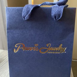 A navy blue shopping bag from Powers Jewelry Designers features gold embossed text and fabric handles, placed on a wooden surface.