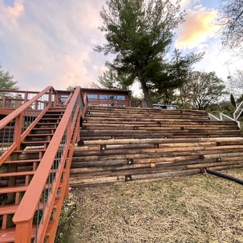 Bob's Digging And Landscaping - the steps leading up to the house - A staircase with metal railings and wooden logs on a hillside, leading up to a building or deck area. The scene includes trees, a partly cloudy sky, and additional outdoor structures with white railings. The ground at the base is covered with grass and dirt, and a black pipe runs along the slope. - Landscaping Near Me - Lodi, Wisconsin