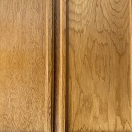 A close-up image of two wooden cabinet doors, showing detailed wood grain patterns and decorative molding at the top edges. The doors are separated by a vertical wooden strip, and the surface appears smooth and finished, highlighting the natural wood texture.
