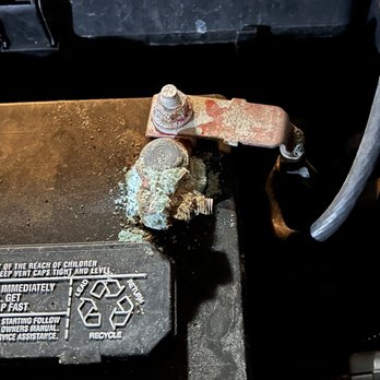 Doc Motor Works - Corroded terminal unaddressed and unmentioned during routine service. - The image depicts a vehicle's battery with a corroded and rusted terminal, showing buildup of corrosion around the connection point. The terminal appears damaged or disconnected, with rust extending onto the connector and surrounding area. The battery is situated in an automotive environment, with visible labels and safety instructions on the battery casing. The corrosion indicates potential electrical issues or the need for maintenance or replacement of the terminal and connector. - Auto Repair Near Me - Plainfield, Illinois