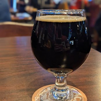 The Globe Pub - Coco Bites Stout Beer - A glass of dark stout beer with a light tan head is placed on a coaster on a wooden table. The background shows a lively bar or pub with blurred patrons, colorful lights, and multiple screens, indicating a sports bar atmosphere. - Sports Bars Near Me - Chicago, Illinois