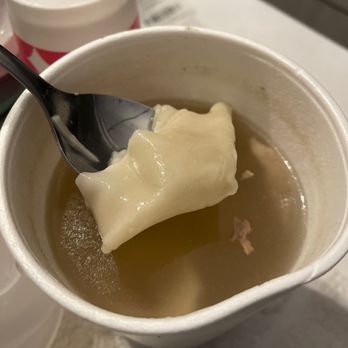 New Dragon - Wonton Soup - A disposable cup filled with clear broth and a single wonton dumpling is shown, with a spoon holding the wonton above the cup. In the background, there are additional containers, possibly of condiments or drinks, on a white surface. - Chinese Near Me - Albuquerque, New Mexico