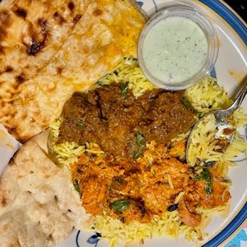 Chit Chaat - Takeout at home: Chicken Tikka Masala & Slow Roasted Beef Curry with Naan garlic + garlic cheese. - A round plate contains yellow rice topped with beef curry and a side of chicken curry. Accompanying the dishes are a piece of naan bread and a small cup of raita. A spoon is placed on the rice, and the plate has a colorful border. - Indian Near Me - Racine, Wisconsin