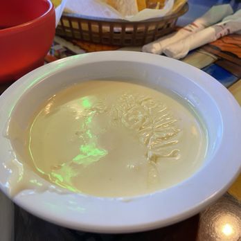 Cinco De Mayo - Queso mmmmm - A white bowl filled with melted queso cheese is positioned on a table, with a basket of tortilla chips lined with paper in the background. A red cup and some napkins are also visible on the table, indicating a casual dining setting, likely at a Mexican restaurant or food event. - Seafood Near Me - Streator, Illinois
