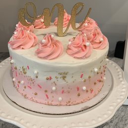Cake - A birthday cake features white and pink frosting with pink rosettes and pearl decorations on top. The cake is adorned with gold and pink sprinkles around the sides and has a gold glittery "one" cake topper. It is placed on a white decorative cake stand on a gray marble surface.