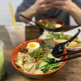 Tonkotsu ramen - A bowl of ramen with vegetables, a boiled egg, and seaweed on a wooden table, with a person taking a photo in the background.