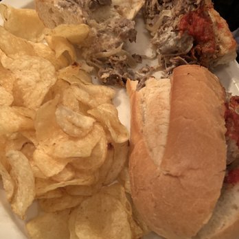 Shenanigans - Cheese Steak - The image shows a white plate holding a cheesesteak sandwich with a bun, potato chips, and slices of meatloaf topped with gravy and tomato sauce. The meatloaf slices are partially cut, revealing the interior, and the chips are piled next to the sandwich. The setting appears to be a dining environment with warm lighting. - Music Venues Near Me - Lake Harmony, Pennsylvania