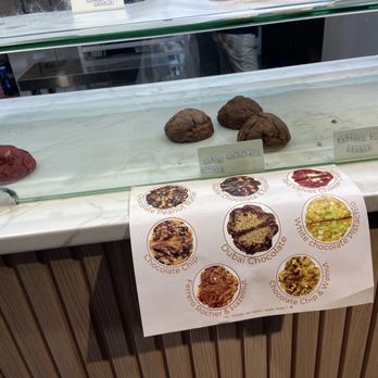 The Sweet Bite - A glass display case contains several cookies, including red velvet and chocolate varieties, with small labels indicating their types. Below the case, a printed menu shows images and names of different cookies such as Dubai Chocolate, White Chocolate Pusacation, and others. The setting appears to be a bakery or cookie shop with a marble countertop and wooden paneling. - Desserts Near Me - Bar Harbor, Maine