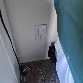 Response Crew - New electrical outlet - The image shows a standard electrical outlet installed on a wall close to a bed. There are cords on the carpeted floor, including a power strip and a device with a remote control. The setting appears to be a bedroom or hotel room. - Handyman Near Me - Scottsdale, Arizona