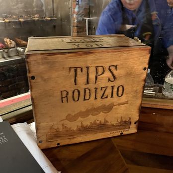 Fernandes steakhouse - a wooden box with the words tips rodizo on it - A wooden tip box with engraved 'TIPS RODIZIO' is placed on a restaurant counter, with a grill and staff visible behind a glass partition. - Portuguese Near Me - Newark, New Jersey