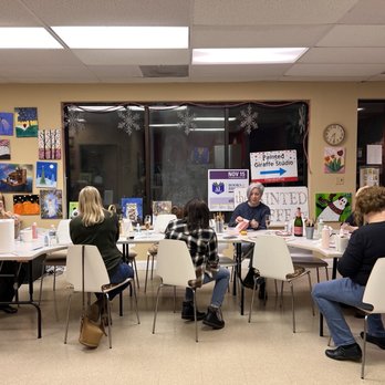 The Painted Giraffe - People are seated around a table in an art studio, engaging in painting activities with art supplies and paintings displayed on the walls. - Art Classes Near Me - Westmont, Illinois