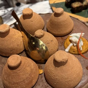 Sukh - Hor Mok - Six chocolate mousse domes with small round tops are arranged on a round platter, accompanied by a spoon and garnishes, in a dining setting. - Thai Near Me - Brooklyn, New York