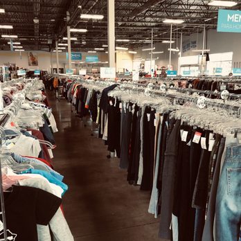 Goodwill Central Texas - Clock Tower - Clothes - The image shows an interior of a thrift store with multiple clothing racks filled with second-hand garments. The racks are organized into sections, with signs overhead indicating areas for children and adults. The store has a high ceiling with exposed metal beams and industrial lighting, and the floor is made of polished concrete. Natural light enters through large windows, illuminating the space. - Used, Vintage & Consignment Near Me - Austin, Texas