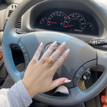 Gloss Nail Spa - The image shows a person's left hand with long, manicured nails painted in a light color, wearing a gold ring with the letter 'M' on the ring finger. The hand is resting on a gray steering wheel of a car, with the dashboard and instrument cluster visible behind, including the speedometer, tachometer, and fuel gauge. The interior of the vehicle appears to be well-maintained, and sunlight is coming through the windshield. - Nail Salons Near Me - Milwaukee, Wisconsin