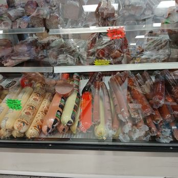 Gastronom - imported meats - The image shows a refrigerated display case in a grocery store, filled with a variety of imported meats, sausages, and processed meat products. The products are wrapped in plastic or casing, with some labels visible. The case has multiple shelves, and the background reflects store lighting and other products. Price tags and labels are attached to some items, indicating their prices and descriptions. - Bakeries Near Me - Arvada, Colorado