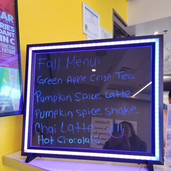 Vibrant Fusion - Fall menu. - A blackboard-style menu with a neon blue border lists fall beverage options such as Green Apple Crisp Tea, Pumpkin Spice Latte, Pumpkin Spice Shake, Chai Latte, and Hot Chocolate. The board is placed on a white surface, and a reflection of a person taking the photo is visible on the glass surface. The background includes a yellow wall and some framed certificates or notices. - Juice Bars & Smoothies Near Me - Hoffman Estates, Illinois